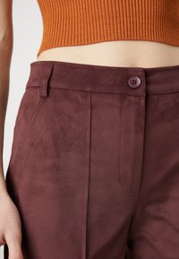 Burgundy faux suede trousers with a smooth texture, featuring a single button closure, side pockets, and tailored fit.