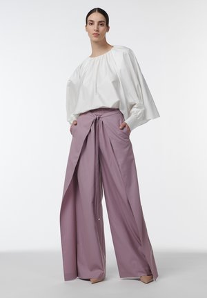 Femme debout portant un pantalon lavande large avec détail nœud devant et une blouse blanche ample à manches longues, mains dans les poches, fond neutre.