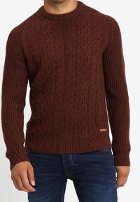 Man i en brun kabelstickad tröja med ribbade muddar och nederkant, kombinerad med mörkblå jeans, visad från hals till höfter.