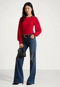 Γυναίκα φορώντας κόκκινο μακρυμάνικο μπλουζάκι με λεπτομέρειες από κουμπιά, μπλε ψηλόμεσες καμπάνες τζιν, μαύρη ζώνη, μαύρα τακούνια και κρατώντας μια μαύρη τσάντα.