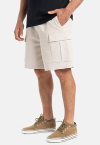 Pantalones cortos cargo beige hechos de una mezcla de algodón, con dos grandes bolsillos laterales y un ajuste relajado, combinados con zapatillas de ante marrón.