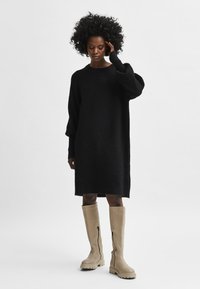 Juoda vilnos mišinys, oversize suknelė ilgomis rankovėmis, apvalia apykakle ir šoninėmis skeltukėmis. Derinama su šviesiai smėlio spalvos iki kelių aukščio auliniais su užtrauktukais.