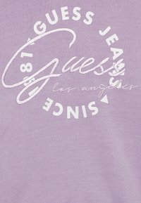 Camiseta de lavanda con texto bordado en blanco: "GUESS," "JEANS," "los ángeles," y "DESDE 1981," con acentos geométricos y textura suave.