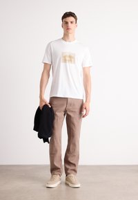 Hvit T-skjorte med grafisk lepedesign; brune bukser; holder en svart genser; beige sneakers; nøytral bakgrunn.