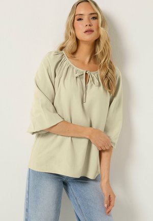 Blonde Frau mit welligem Haar trägt eine lockere, hellgrüne Bluse mit Bindeband am Ausschnitt und hellblaue Jeans und posiert vor einer weißen Wand.