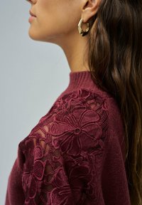 Perfil lateral de una mujer con cabello largo y oscuro, vistiendo un suéter burdeos con bordado de encaje floral en el hombro y un pendiente de aro dorado.