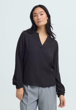 Mujer vistiendo una blusa negra suelta con cuello en V y mangas largas abullonadas, y pantalones plisados de color gris claro, de pie frente a un fondo claro y sencillo.