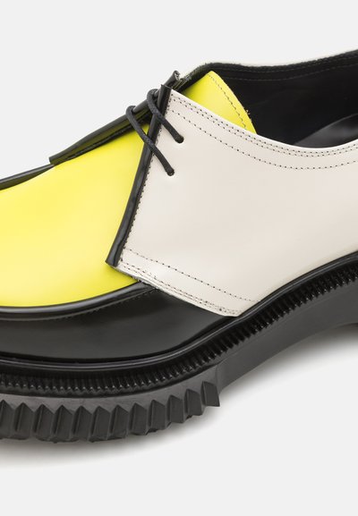 Zweifarbiger Lederschuh mit gelbem und weißem Obermaterial, schwarzer Sohle mit strukturiertem Profil und schwarzen Schnürsenkeln. Rundliche Zehenform.