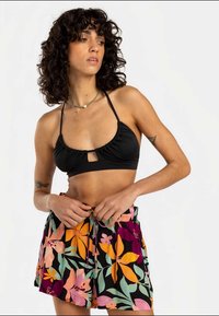 Kvinne med krøllete hår iført en svart bikini-topp og fargerike blomstrete shorts som justerer livet, ser mot venstre mot en ensfarget bakgrunn.