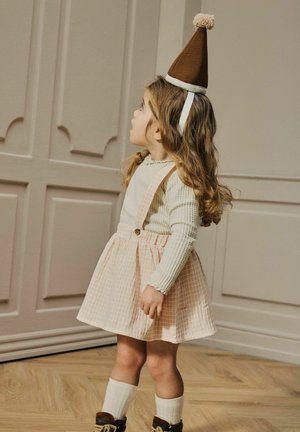 Niña pequeña con un sombrero de fiesta marrón, suéter color crema, falda con tirantes a cuadros rosas, calcetines blancos y botas marrones, de pie en el interior.