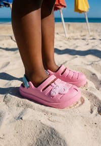Jambes d’enfant avec des chaussures slip-on roses ornées d’un hippopotame de dessin animé, debout sur une plage de sable avec des parasols colorés en arrière-plan.