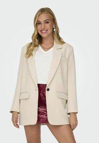 Blazer crema con bottone nero, due tasche frontali e vestibilità sartoriale, indossato sopra un top bianco a V e una gonna color borgogna con paillettes.