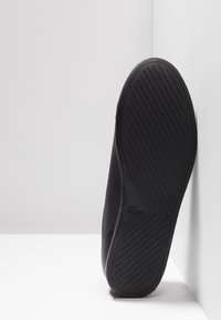 Semelle de basket noire avec un motif de bande de roulement diagonal reposant verticalement contre un mur d'angle blanc et un sol blanc.