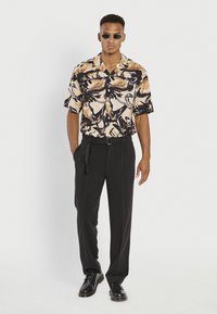 Chemise à manches courtes, avec un motif abstrait beige et noir, associée à un pantalon noir et des chaussures à lacets noires. Les accessoires incluent des lunettes de soleil sombres.