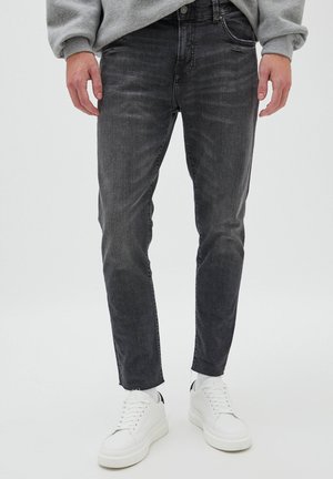 Homme portant un jean noir délavé slim fit et des baskets blanches, debout devant un fond clair uni.