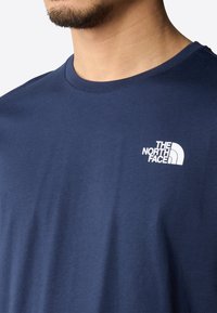 Námořnická modrá bavlněná tričko s kulatým výstřihem, s bílým logem na levé části hrudi. Hladká textura a neformální design.