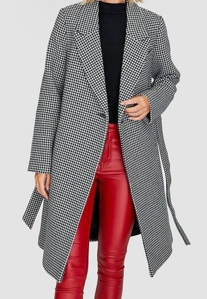 Femme portant un manteau pied-de-poule noir et blanc par-dessus un top noir et un pantalon en cuir rouge, debout devant un fond uni.