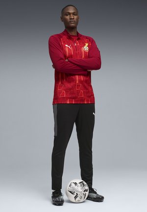 Hombre de pie con los brazos cruzados, vestido con una chaqueta de fútbol de Ghana roja, pantalones negros y tacos, con un balón de fútbol en blanco y negro a sus pies.