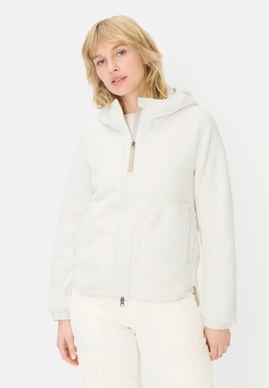 Młoda kobieta o blond włosach, ubrana w białą polarową bluzę na zamek z kapturem i jasne spodnie, stojąca na jednolitym tle.