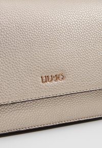 Gros plan sur une surface en cuir texturé beige avec des lettres de logo en métal doré "LIU·JO" et des coutures visibles sur les bords.