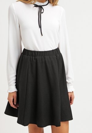 Vrouw draagt een witte top met lange mouwen met een zwarte lintstrik en een zwarte A-lijn rok met hoge taille en elastische tailleband.