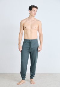 Graue Jogginghosen mit einem Kordelzug, schmal zulaufendem Bein und weicher Textur, getragen von einem männlichen Model ohne Hemd.