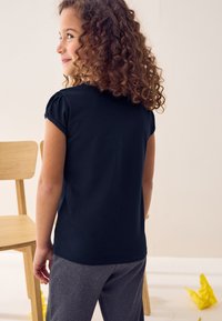 Schwarzes kurzärmeliges Hemd mit Raffung, kombiniert mit grauen Hosen. Der Hintergrund zeigt Möbel aus hellfarbenem Holz und zerknülltes gelbes Papier.