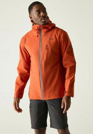 Mann trägt eine leuchtend orangefarbene Outdoor-Jacke mit Kapuze und schwarze Shorts, schaut nach links vor einem einfarbigen hellen Hintergrund.
