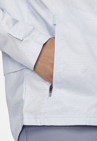 Mano insertada en un bolsillo lateral parcialmente desabrochado de una chaqueta de patrón de cuadrícula azul claro con una manga acampanada.
