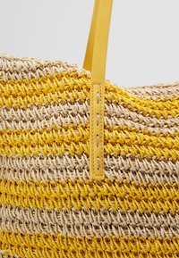 Gros plan d'un sac tissé avec des rayures alternées jaune et beige naturel, et une sangle en cuir jaune fixée avec une couture visible.