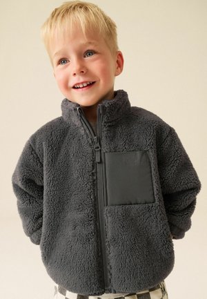 Junger blonder Junge lächelt, trägt eine dunkelgraue Fleecejacke mit Frontreißverschluss und einer quadratischen Brusttasche.