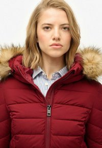 Chaqueta tipo puffer burdeos con capucha forrada de piel, costuras horizontales y cremallera frontal. El cuello interior presenta un detalle de tela a rayas.