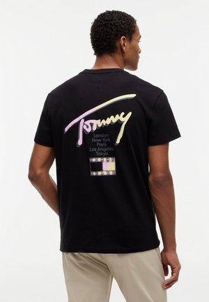 Άνδρας που φοράει μαύρο μπλουζάκι με παστέλ γράμματα "Tommy", ονόματα πόλεων και ένα μικρό καρό λογότυπο στο πίσω μέρος, συνδυασμένο με μπεζ παντελόνι.