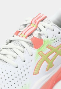 Λευκό αθλητικό παπούτσι με τρυπητό άνω μέρος, ροζ και πράσινες λεπτομέρειες, και υφή στη σόλα. Διαθέτει κορδόνια και δαχτυλίδι στη φτέρνα για εύκολη χρήση.