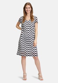 Kurzärmliges Kleid im schwarz-weißen Zickzackmuster, weicher Stoff, V-Ausschnitt, A-Linien-Silhouette, knielang, kombiniert mit weißen Absatzsandalen.