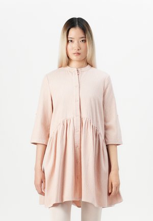 Jeune femme avec des cheveux ombrés raides portant une robe boutonnée rose pâle ample à manches trois-quarts sur fond blanc.