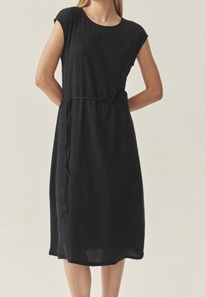 Femme portant une robe noire texturée jusqu'au genou, avec un col rond, des manches courtes style capes et une fine ceinture nouée à la taille.