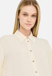 Helle Creme-Bluse mit Kragen, vorderen Knöpfen und glatter Stofftextur. Accessoires mit langen, silbernen, hängenden Ohrringen.
