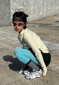 Jauna persona saulesbrillēs, krēmkrāsas teksturētu garroku krekliņā, zilās legingās, svītrainās zeķēs un baltiem sporta apaviem, tup uz akmens bruģa.