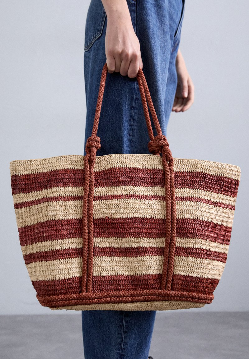 Torebka z tkanego słomy z czerwonymi i kremowymi paskami, z dwiema plecionymi uchwytami ze sznurka i naturalną teksturą. Lekko usztywniony kształt.