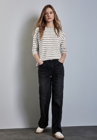 Gestreepte pullovertrui in crème en grijs, gecombineerd met zwarte wijde jeans. Model draagt witte sneakers. Eenvoudig, casual outfitontwerp.