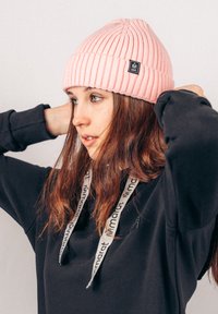 Roze geribbelde gebreide beanie met een zwart label, gedragen met een zwarte hoodie met witte trekkoorden, die een gladde textuur vertoont.