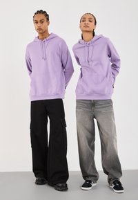 Felpe viola con tasche frontali, indossate con pantaloni cargo neri e jeans grigi sbiaditi. Sneaker con accenti neri e bianchi completano l'outfit.