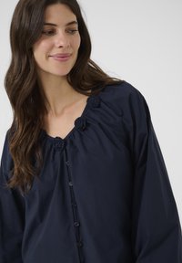 Blouse bleu marine avec un décolleté en V, des détails froncés et des roses en tissu décoratives au niveau du cou. Présente des boutons sur le devant.