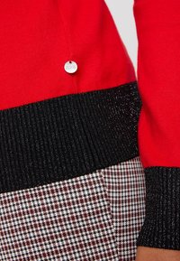 Czerwona bluza z czarnym, błyszczącym ściągaczem, w połączeniu z wzorzystym dołem w kratę w kolorze burgundowym, czarnym i białym. Widoczny okrągły detal z logo.