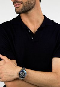 Polo shirt da uomo nero con maniche corte, caratterizzato da un colletto classico e un orologio argento con ghiera blu su un bracciale in acciaio inossidabile.