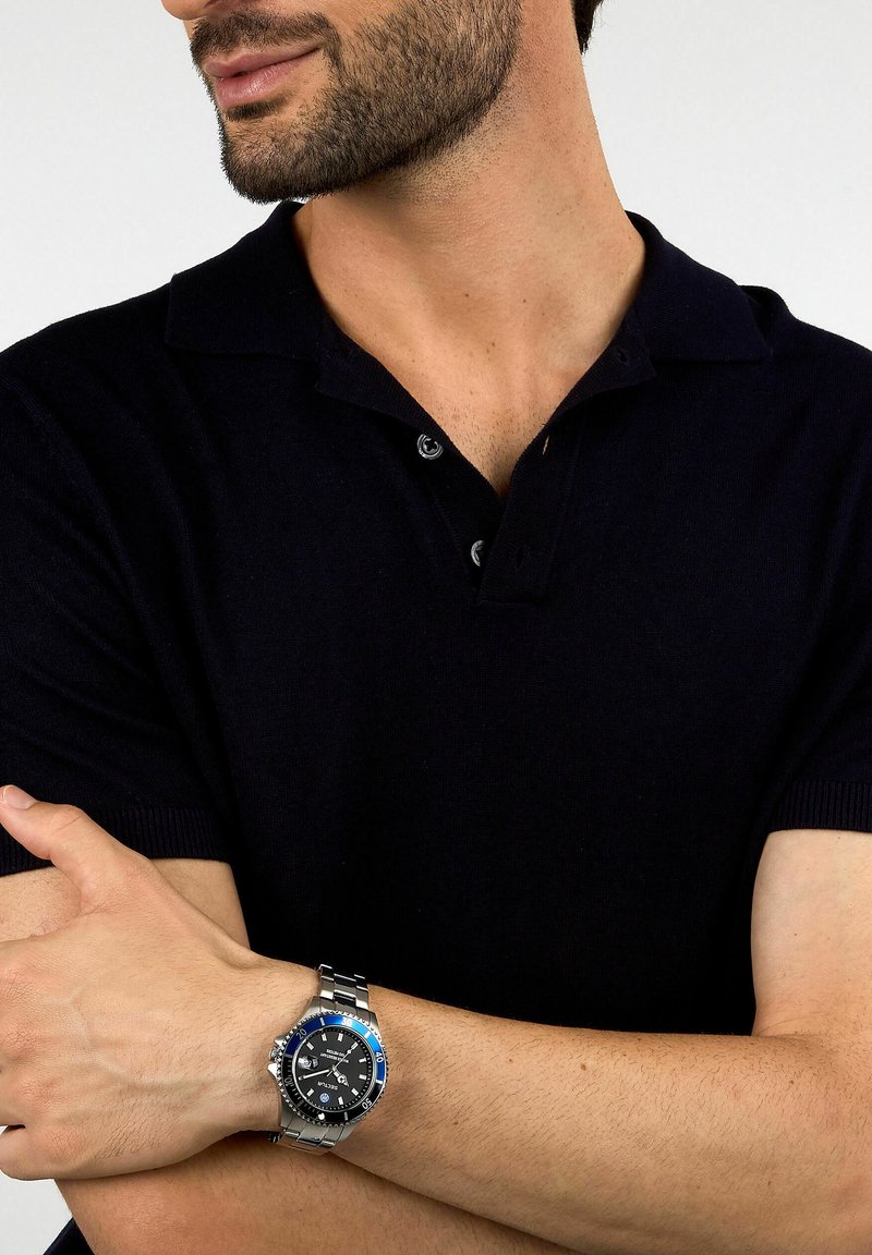 Polo shirt da uomo nero con maniche corte, caratterizzato da un colletto classico e un orologio argento con ghiera blu su un bracciale in acciaio inossidabile.