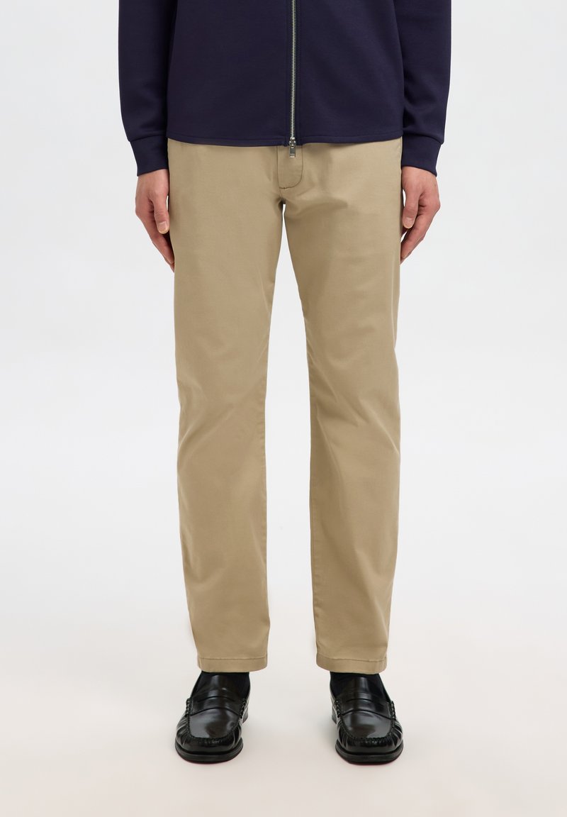 Mann, der beige, gerade geschnittene Hosen, schwarze Loafers und eine marineblaue Zip-up-Jacke trägt, steht vor einem hellen Hintergrund.