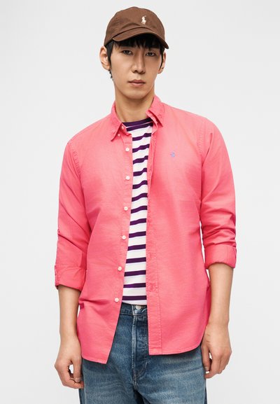 Νέος άνδρας που φοράει καφέ καπέλο, φωτεινό ροζ πουκάμισο με κουμπιά πάνω από λευκό και μωβ ριγέ T-shirt και μπλε τζιν, στέκεται μπροστά σε απλό φόντο.