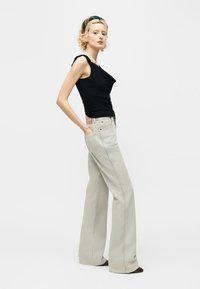 Žena s krátkymi kučeravými vlasmi, oblečená v čiernom tielku bez rukávov, béžových širokých džínsoch, perlovom náhrdelníku a vzorovanej čelenke, stojaca s rukami v vreckách.